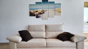 A beige sofa with two dark cushions, a wall-mounted artwork depicting a beach scene, and a window with blinds. - GERONA sea view apartments (Benidorm)