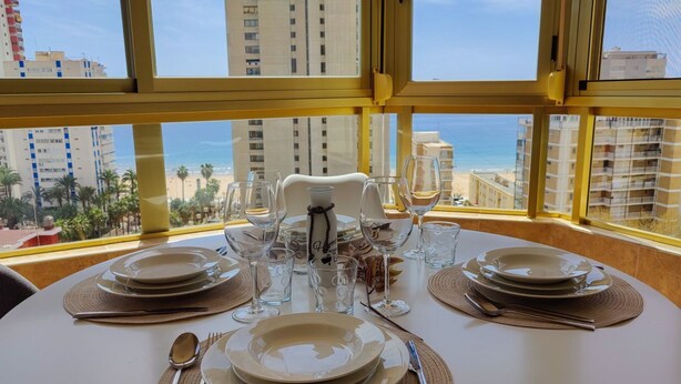 A dining table set for four with a view of the beach and buildings through large windows. - GERONA sea view apartments (Benidorm)