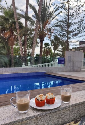A poolside area with a clear view of the pool, a glass of coffee, and two cupcakes on a plate.