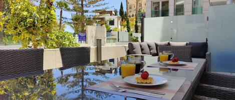 A dining area with a glass table, wicker furniture, and a view of buildings and trees.