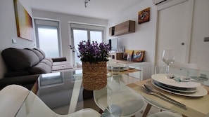 A modern living room with a glass dining table, white furniture, and a vase of purple flowers.