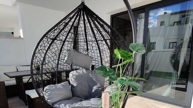 Un espace de vie moderne avec un fauteuil suspendu, une petite table ronde sur laquelle se trouvent une bouteille et des verres, et une plante en pot.