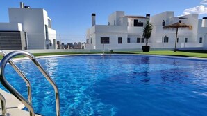 Une piscine entourée de rampes métalliques, bordée de bâtiments modernes blancs et surplombée par un ciel bleu clair.