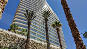 A tall, modern hotel with a curved facade, palm trees in front, and a stone base. - SUNSET CLIFFS sea & sky apartments (Benidorm)