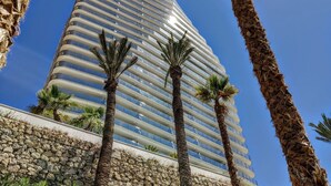 A tall, modern hotel with a curved facade, palm trees in front, and a stone base. - SUNSET CLIFFS sea view apartments (Benidorm)