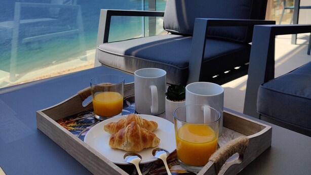 Ein Frühstücksbrett mit Croissants, Orangensaft und Kaffee auf einem Balkon mit Blick aufs Meer.