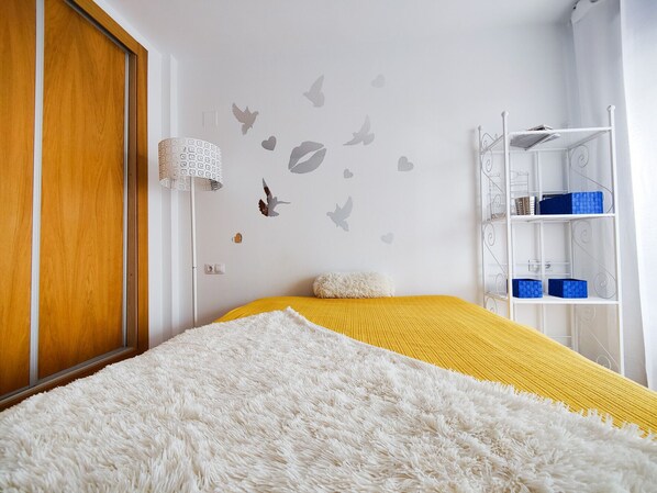A bedroom with a wooden door, a white fluffy rug, a yellow bedspread, a white fluffy pillow, a white metal shelf with blue storage boxes, and wall decals of birds and hearts.
