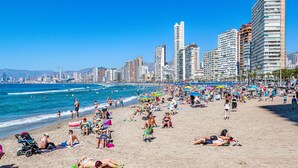 Une plage bondée où les gens se prélassent au soleil et se baignent, entourée de grands immeubles et surplombée d’un ciel bleu clair.