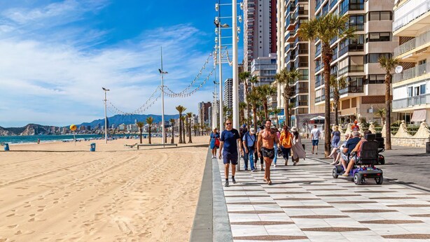 Un passeig marítim al costat d'una platja, amb un paviment d'escacs, palmeres i edificis alts al fons.