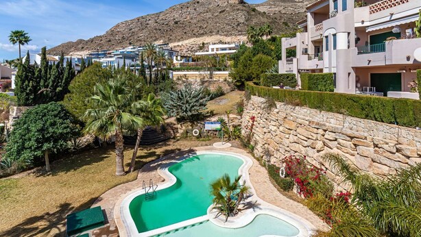 A swimming pool with a wavy design, surrounded by stone walls and lush greenery. - Jardin Sweet Apartment (Finestrat)