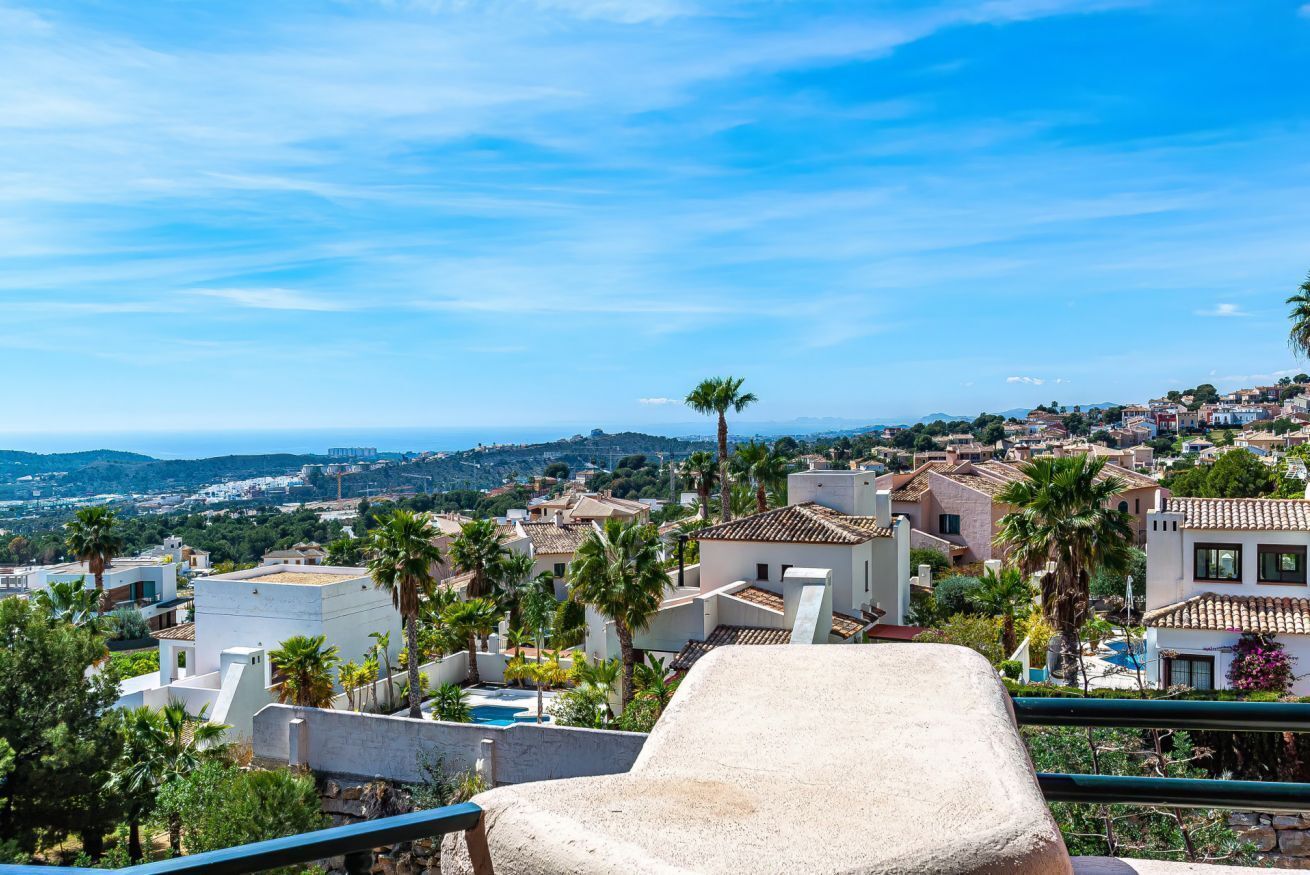 Una terraza con banca de piedra con vistas a un pueblo costero de arquitectura mediterránea, palmeras y cielo azul despejado.