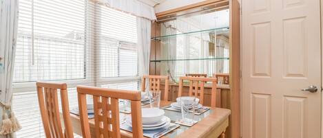 Un área de comedor con una mesa de madera, sillas y una mesa con cubierta de vidrio. Se observa una puerta y una ventana con persianas.