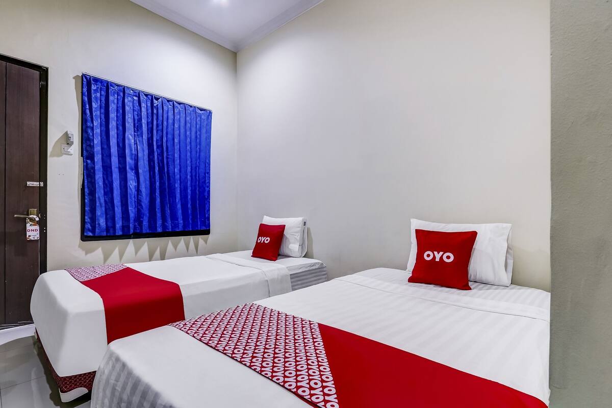 Quarto de hotel com duas camas, uma cortina azul e um travesseiro vermelho com a inscrição 'OYO'.