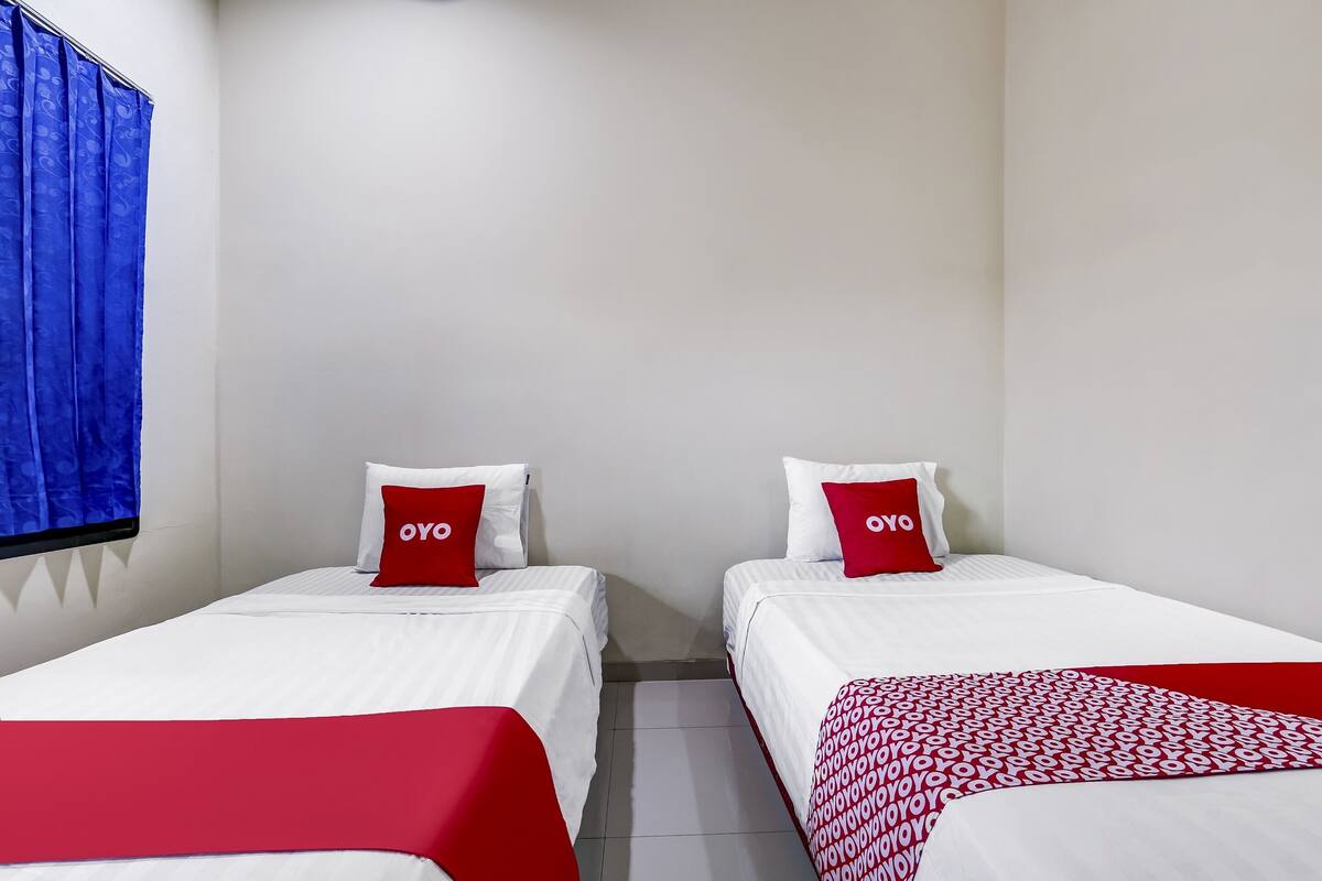 Quarto de hotel com duas camas, cada uma com roupa de cama branca e detalhes vermelhos, incluindo almofadas e um cobertor.