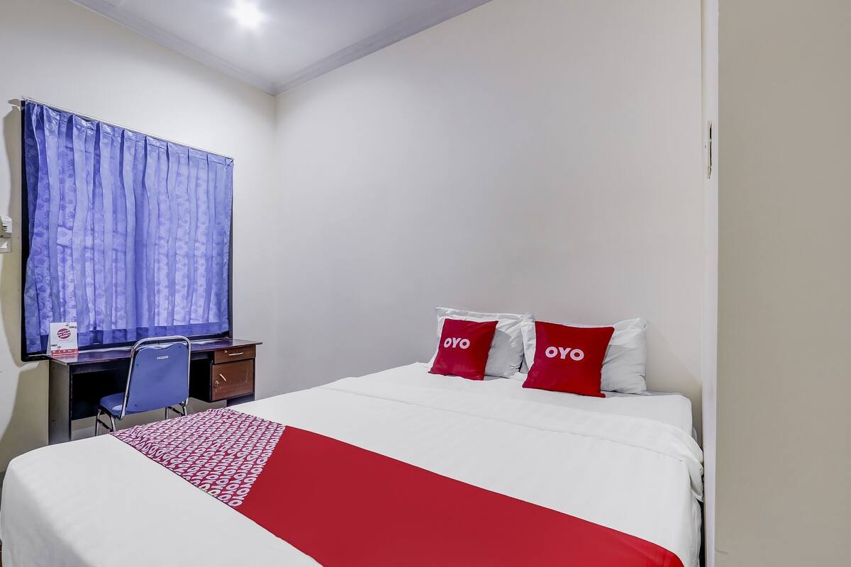 Quarto de hotel com cama, escrivaninha, cadeira e janela com cortinas.