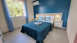 A hotel room with two beds, a blue wall, an air conditioning unit, and a window with curtains. - Paradis Bleu Residence (Petit-Bourg)