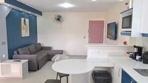 A compact living space with a sofa, a small round table, a kitchenette, and a TV mounted on the wall. - Paradis Bleu Residence (Petit-Bourg)