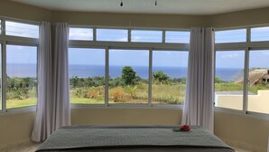 A bedroom with a bed, a ceiling fan, and large windows offering a view of the ocean and greenery. - Cabrera oceanview villa (Cabrera)