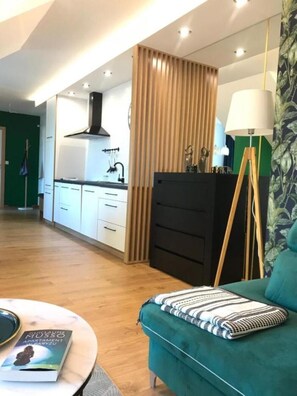 Un salon moderne avec un canapé de couleur turquoise, une table basse sur laquelle repose un livre, et un coin cuisine doté de meubles blancs et d’une hotte noire.