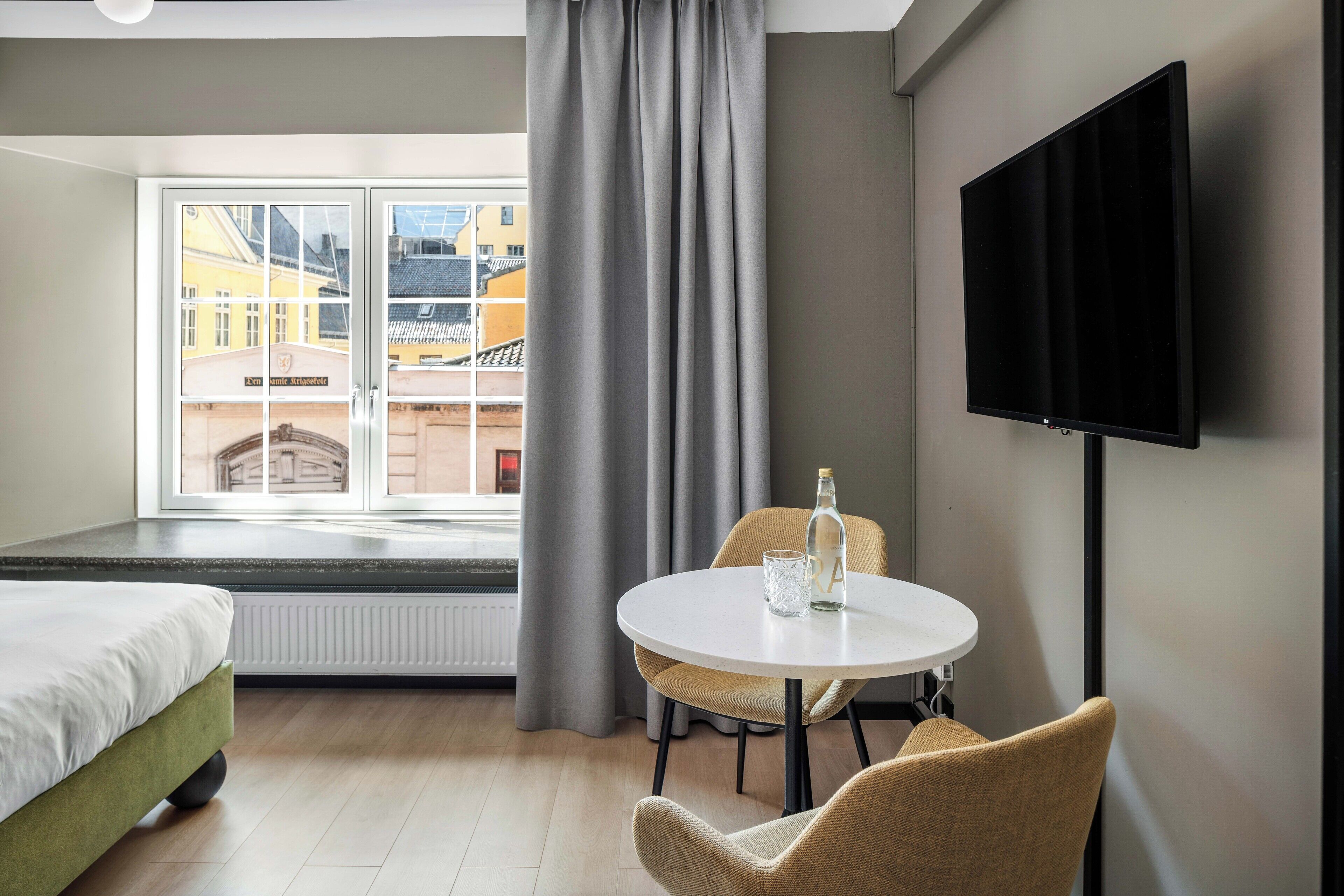 Quarto de hotel com cama, mesa redonda e uma cadeira, televisão na parede e janela com cortinas.