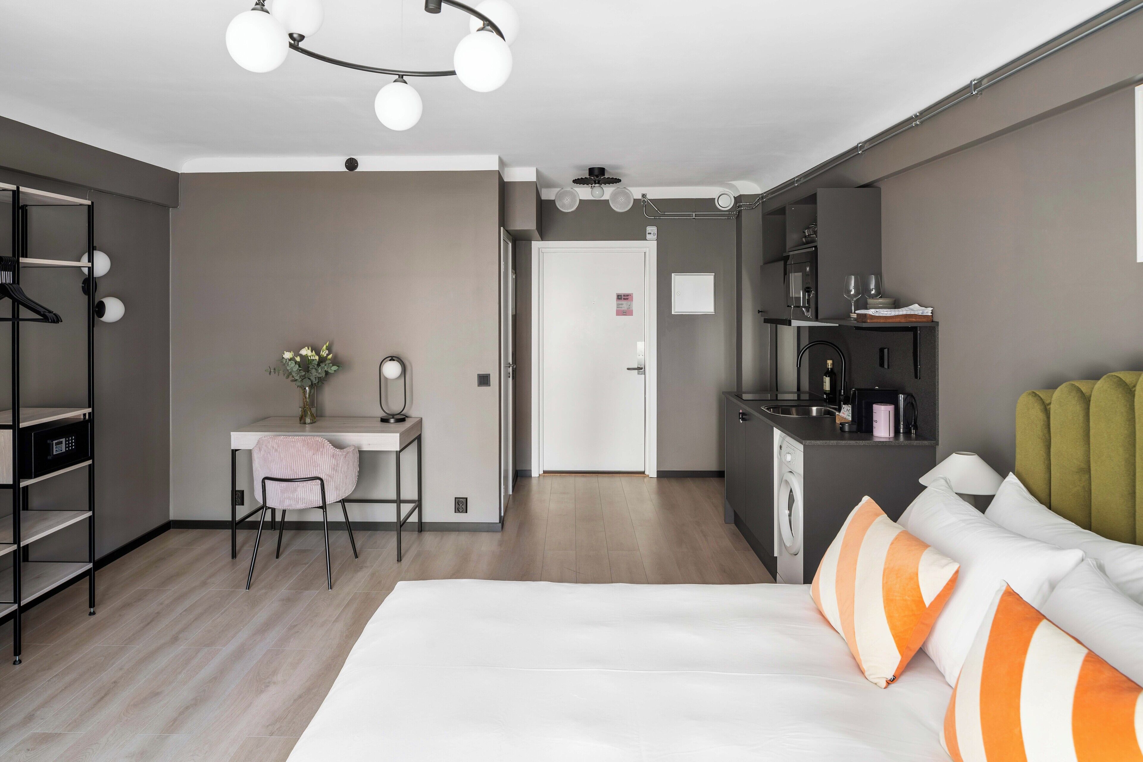 A modern hotel room with a bed, a desk, a chair, a wardrobe, a mirror, and a kitchenette.