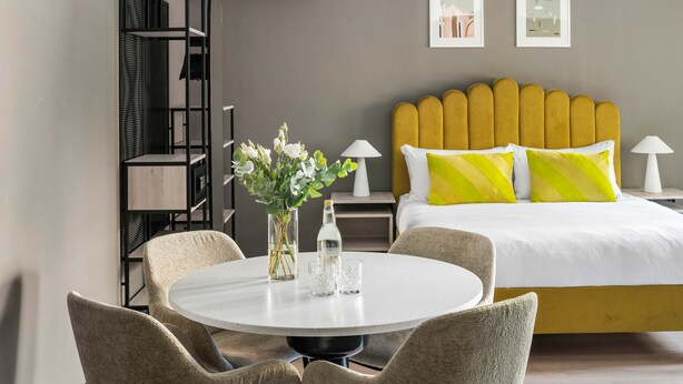 Een moderne hotelkamer met een ronde eettafel, twee stoelen, een bed met een geel hoofdeinde en twee nachtkastjes met lampen.