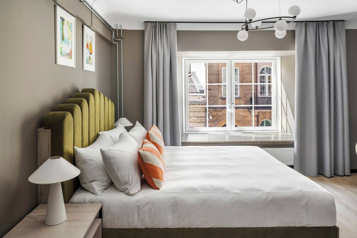 Une chambre moderne avec un grand lit, une lampe de chevet et une vue sur un immeuble par la fenêtre.