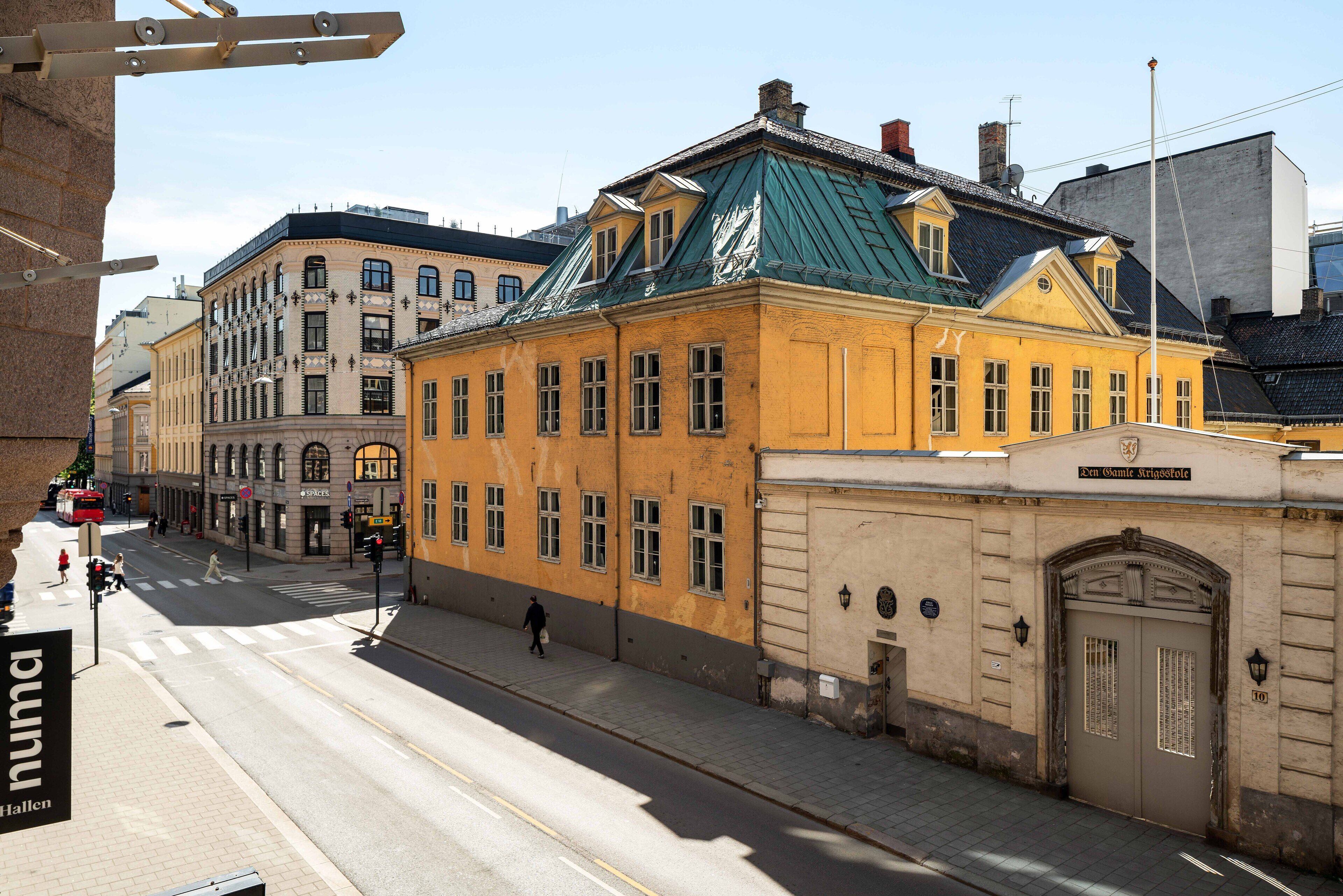 A street view with a yellow building with a green roof, a stone building with a sign, and a pedestrian crossing.