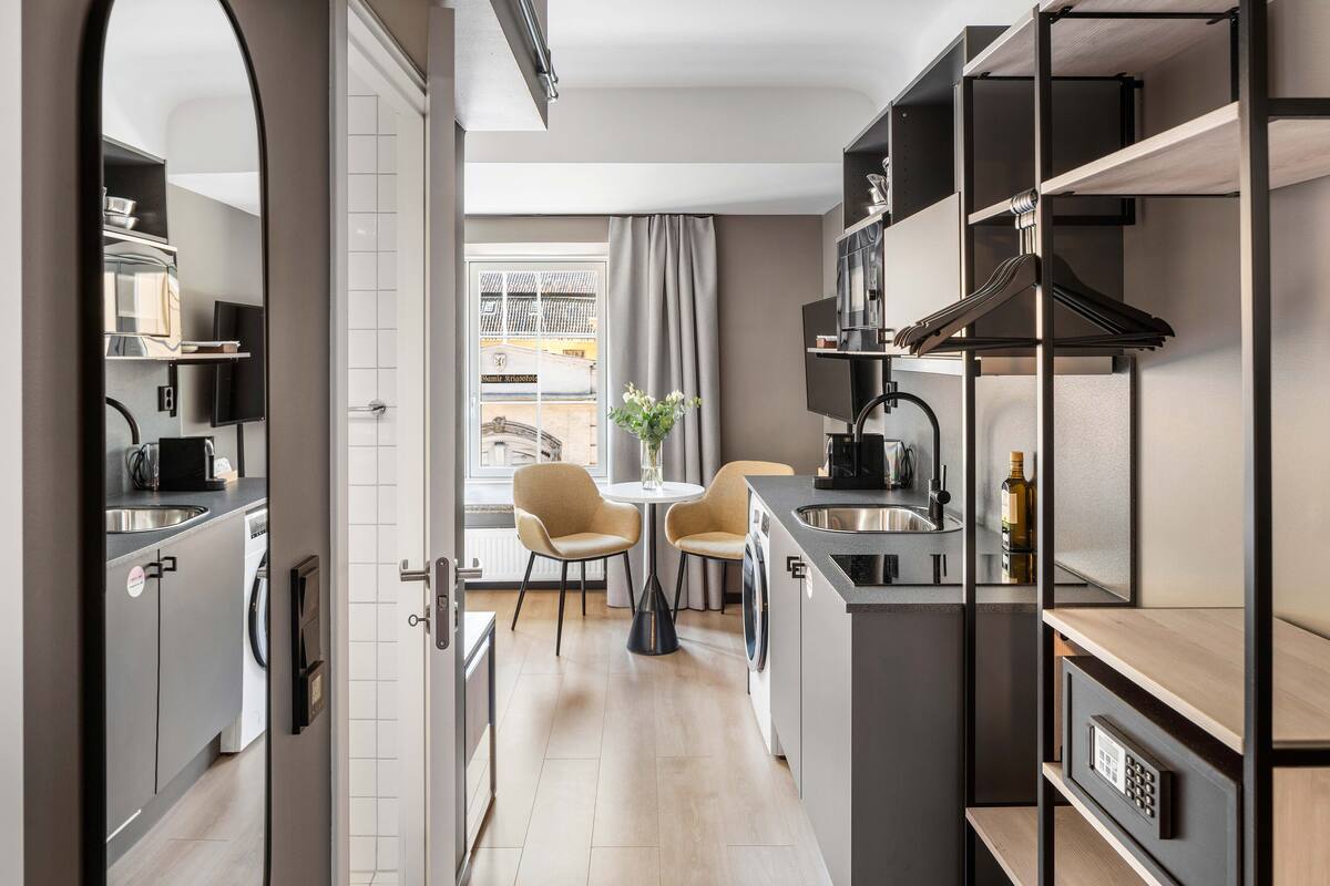 Une cuisine compacte dotée d’appareils électroménagers modernes, un coin repas avec deux chaises et une vue sur la rue grâce à une grande fenêtre.