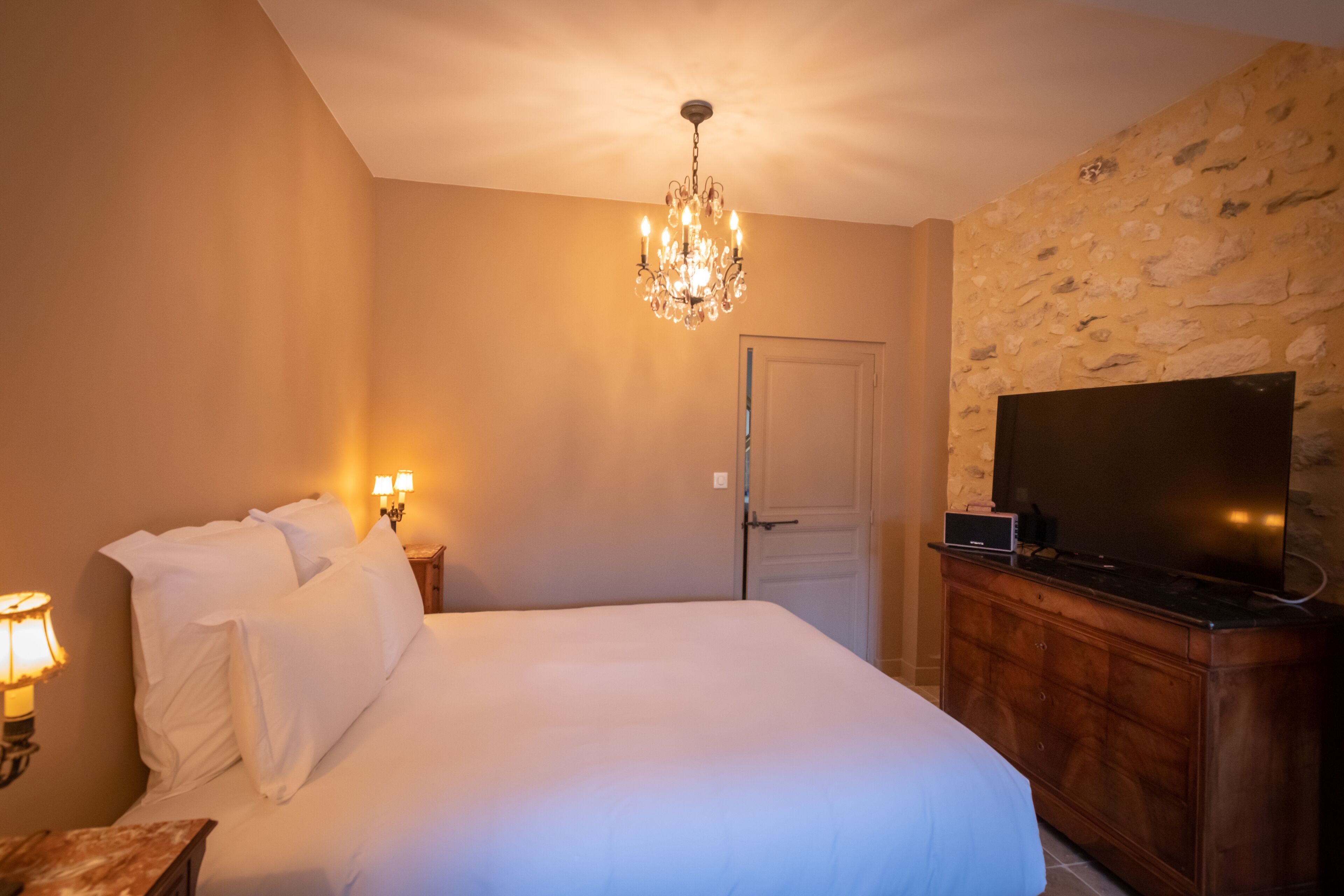 A bedroom with a large bed, a chandelier, a television on a wooden cabinet, and a stone wall.