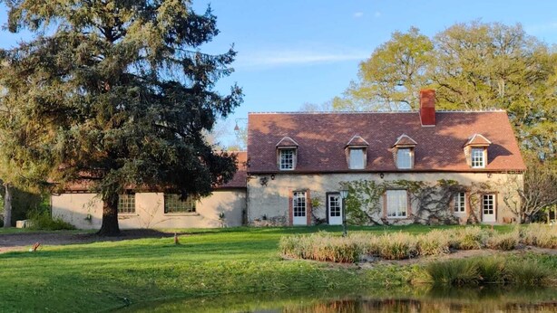 Une maison à deux étages avec un toit recouvert de tuiles rouges, un étang au premier plan et un grand arbre à gauche.