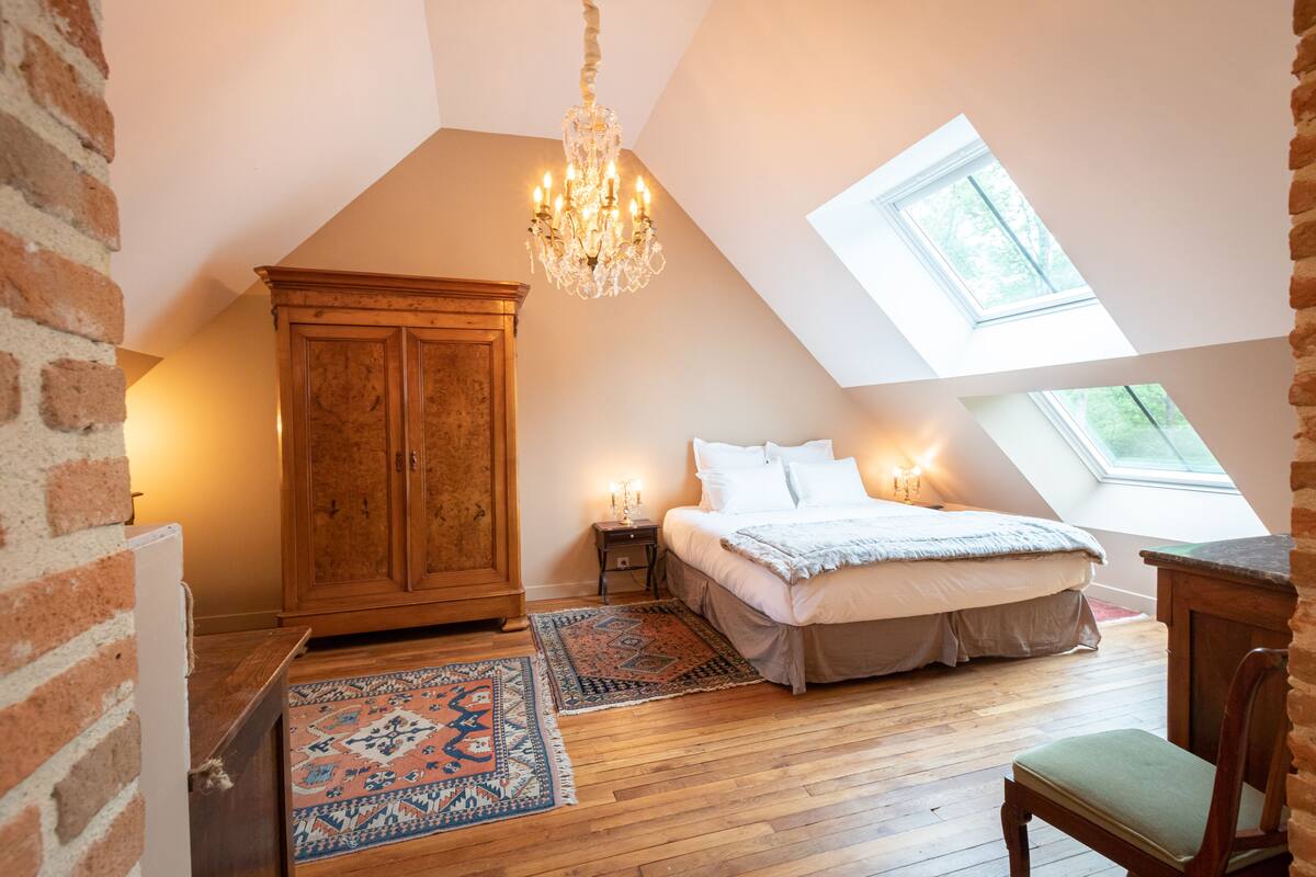 Ein Schlafzimmer mit schrägen Wänden, einer Deckenleuchte, einem Bett, einem Nachttisch, einem Holzschrank und einem Oberlicht.