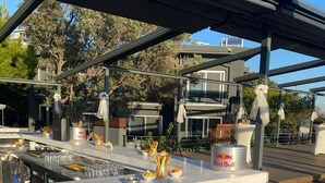 A modern outdoor bar area with a wooden bar counter, white stools, and a Red Bull branded cooler. - Denizati Plus Hotel (Istanbul)