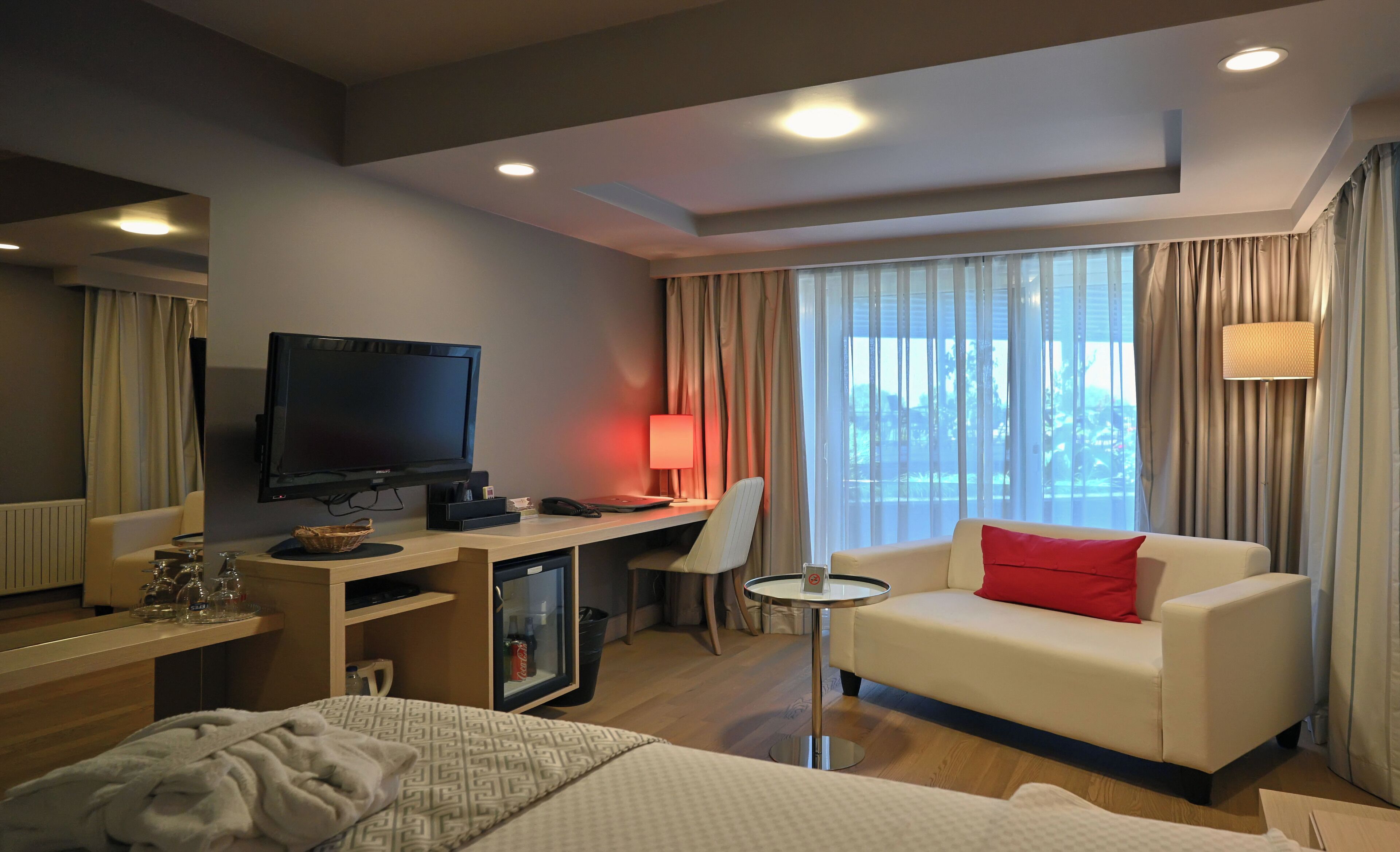 Une chambre d’hôtel avec un lit, un bureau avec un ordinateur, une télévision, un canapé avec un coussin rouge et une fenêtre avec des rideaux.