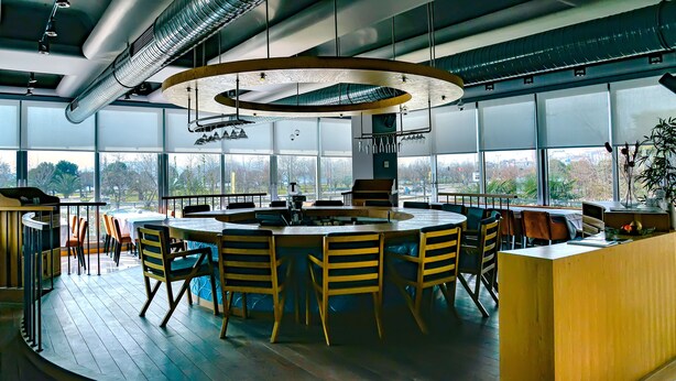 A modern dining area with a central round table surrounded by chairs, large windows offering a view of the outdoors, and a ceiling with exposed ductwork. - Denizati Plus Hotel (Istanbul)