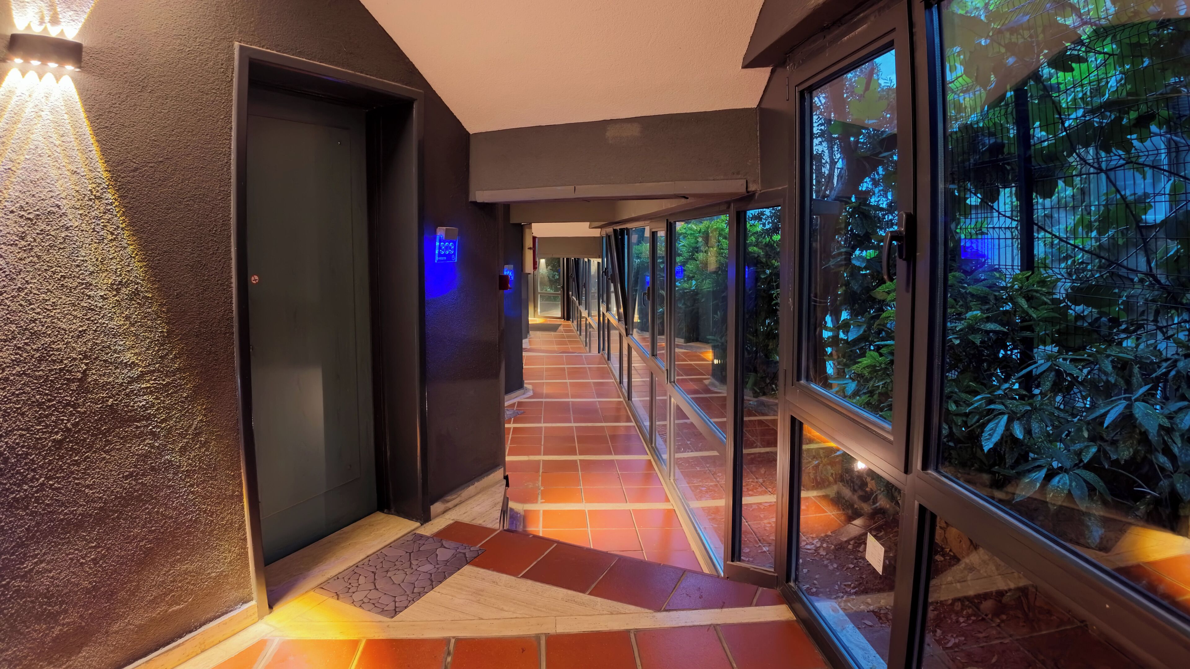 A corridor with a tiled floor leading to a door, glass walls, and a view of greenery outside.