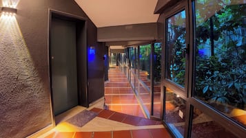 A corridor with a tiled floor leading to a door, glass walls, and a view of greenery outside.