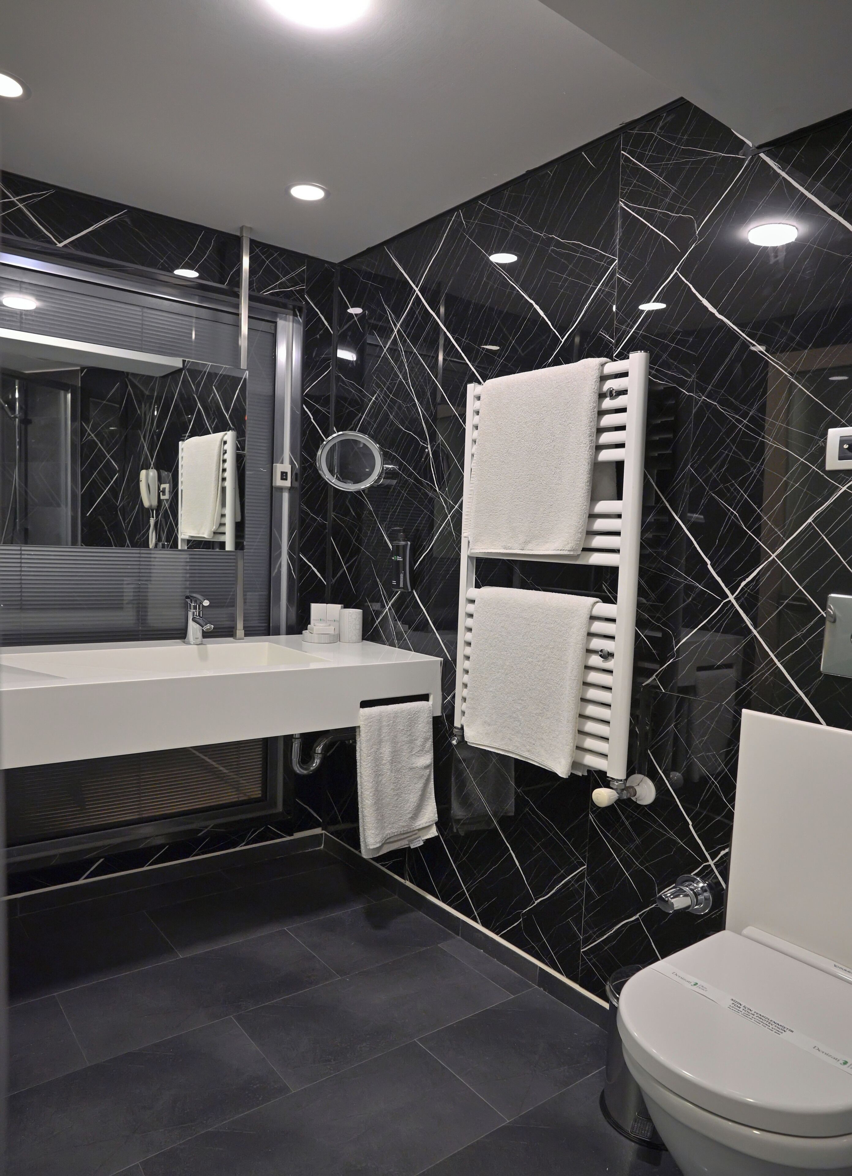 Une salle de bain moderne avec un mur en marbre noir, un lavabo blanc, un miroir, un porte-serviettes et des toilettes.