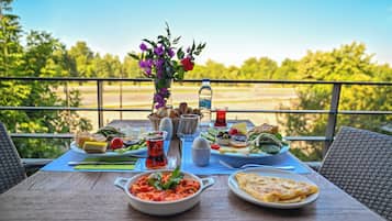 A table set with a variety of dishes, a bottle of water, and a vase of flowers on a balcony overlooking a green landscape.