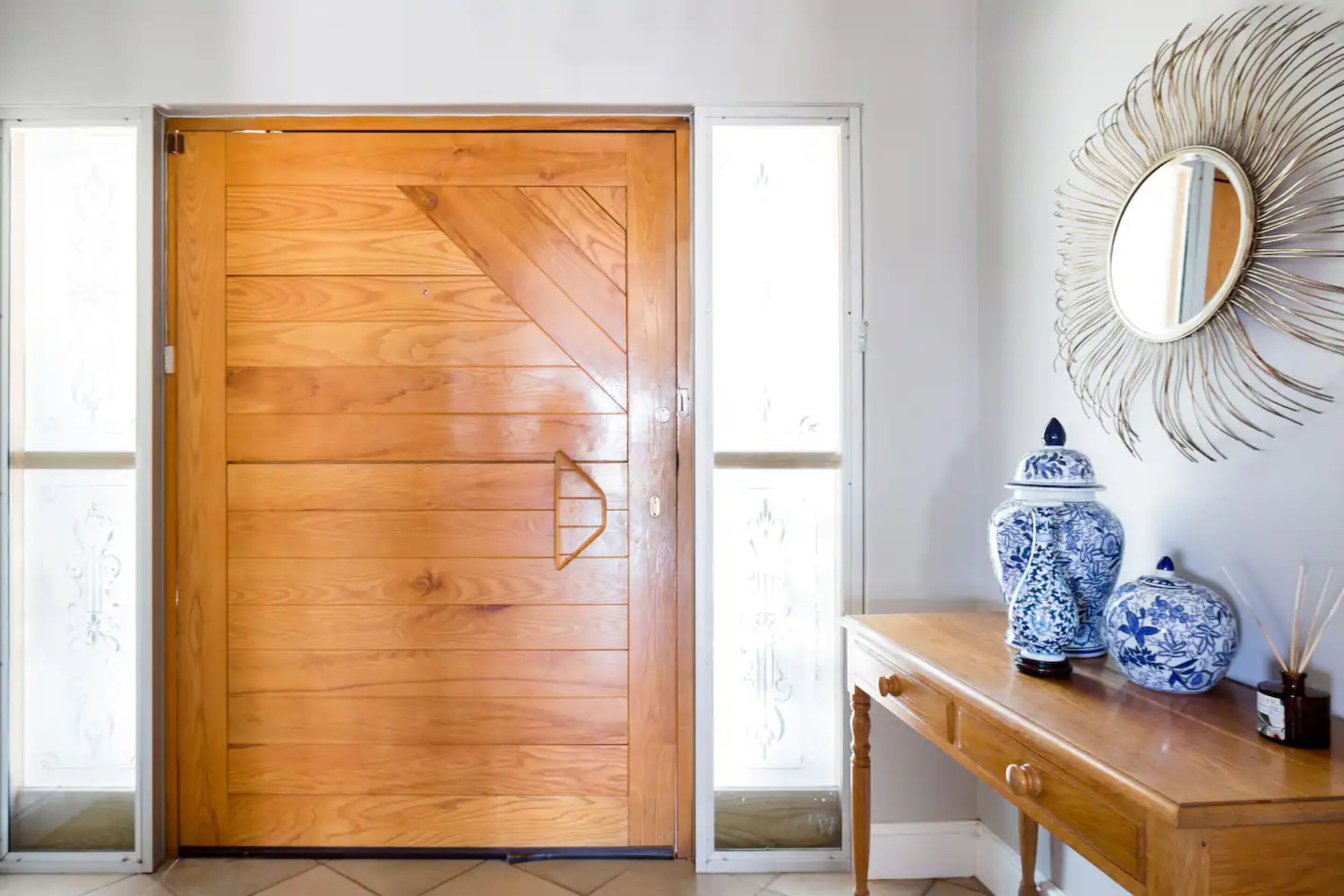 A wooden door with a geometric pattern, a mirror with a sunburst frame, and two blue and white vases on a wooden console table.