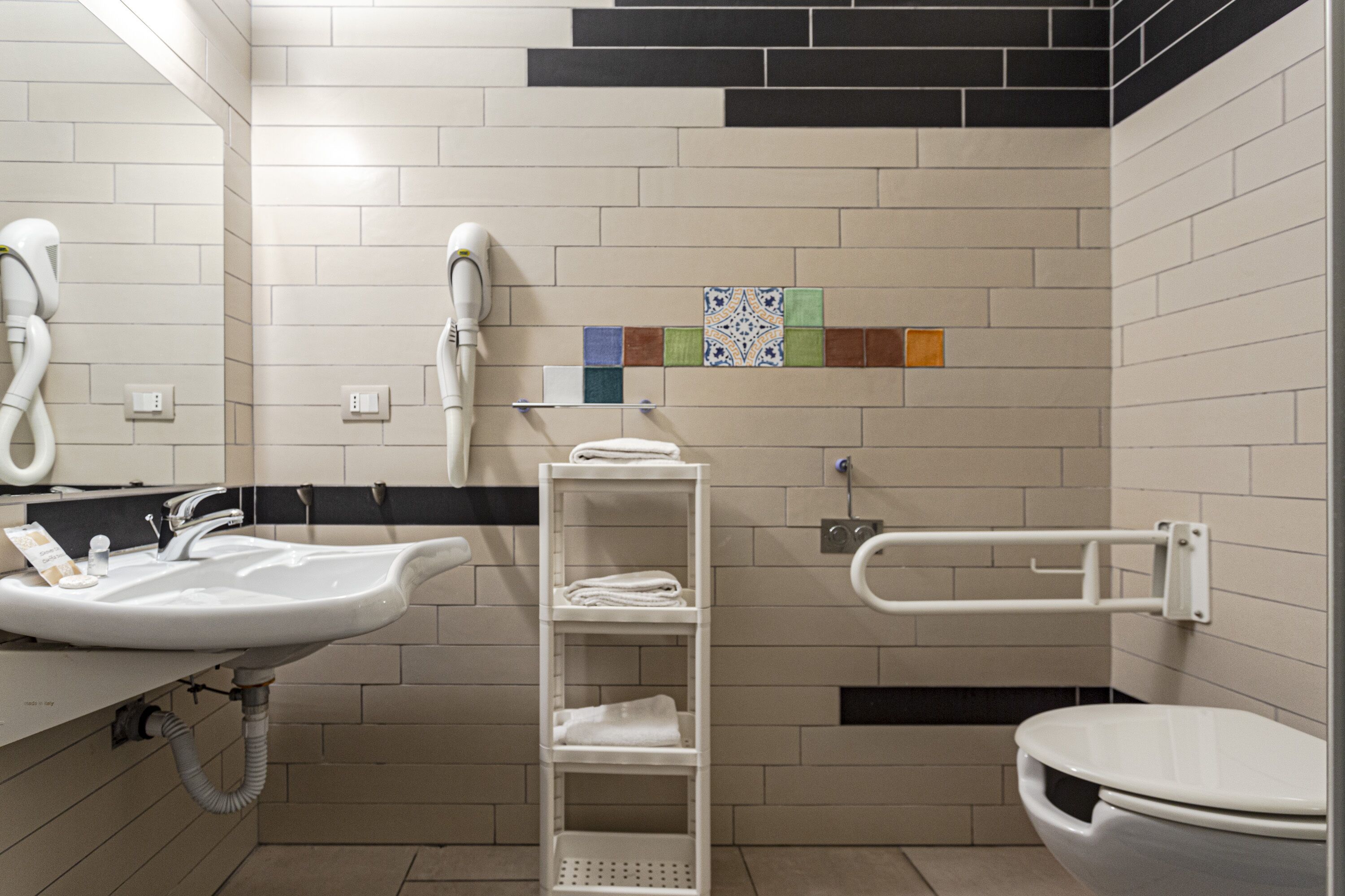 Ein modernes Badezimmer mit Waschbecken, Toilette und Handtrockner. Eine Ablagefläche mit Handtüchern und einem kleinen dekorativen Fliesenmuster an der Wand.