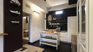 Una cocina moderna con armarios blancos, encimera de madera y salpicadero negro. Hay tres lámparas colgantes, una cafetera y un estante pequeño con adornos.
