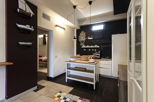 Una cocina moderna con armarios blancos, encimera de madera y salpicadero negro. Hay tres lámparas colgantes, una cafetera y un estante pequeño con adornos.