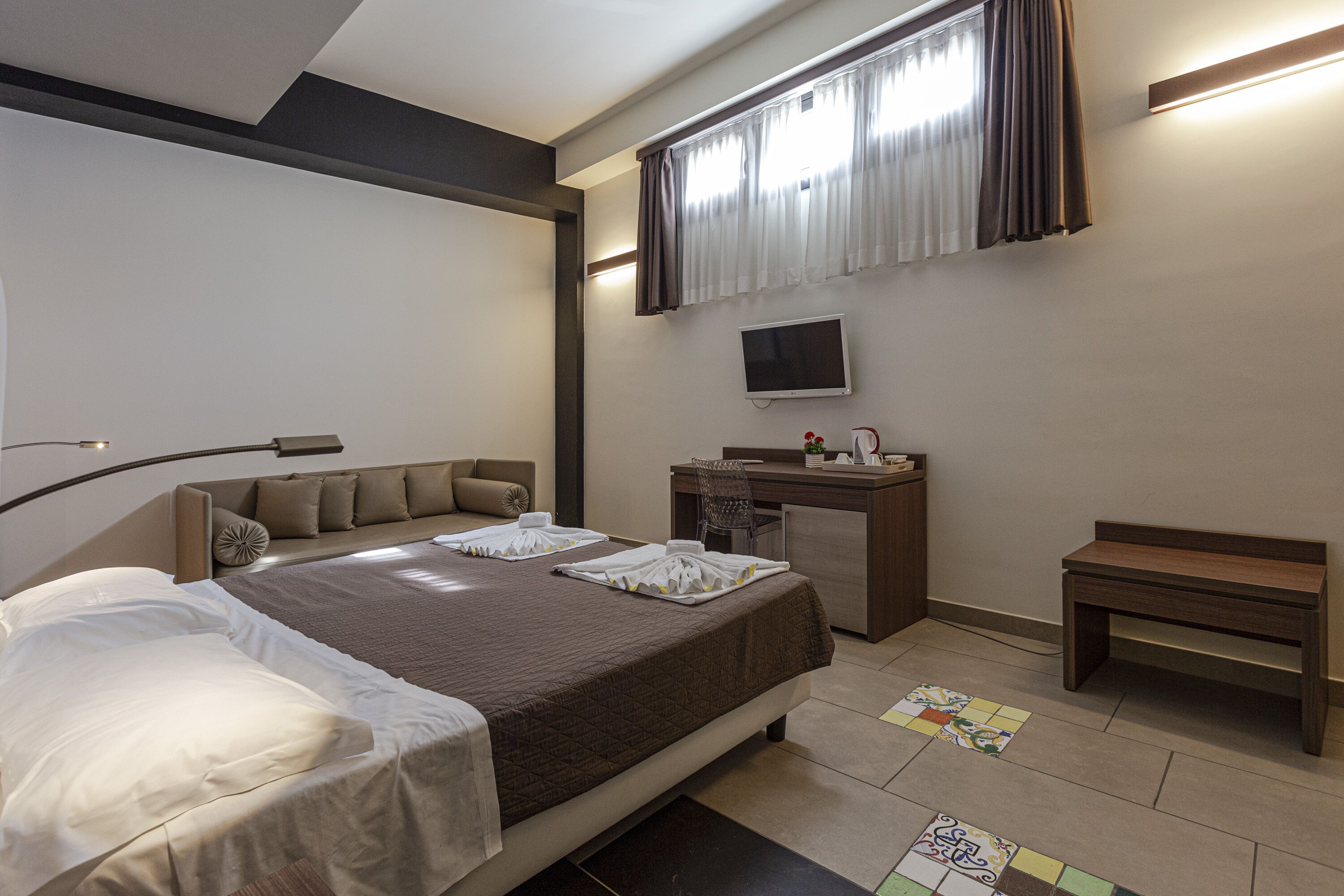 A hotel room with a bed, a desk, a chair, a small table, a TV, and a window with curtains.