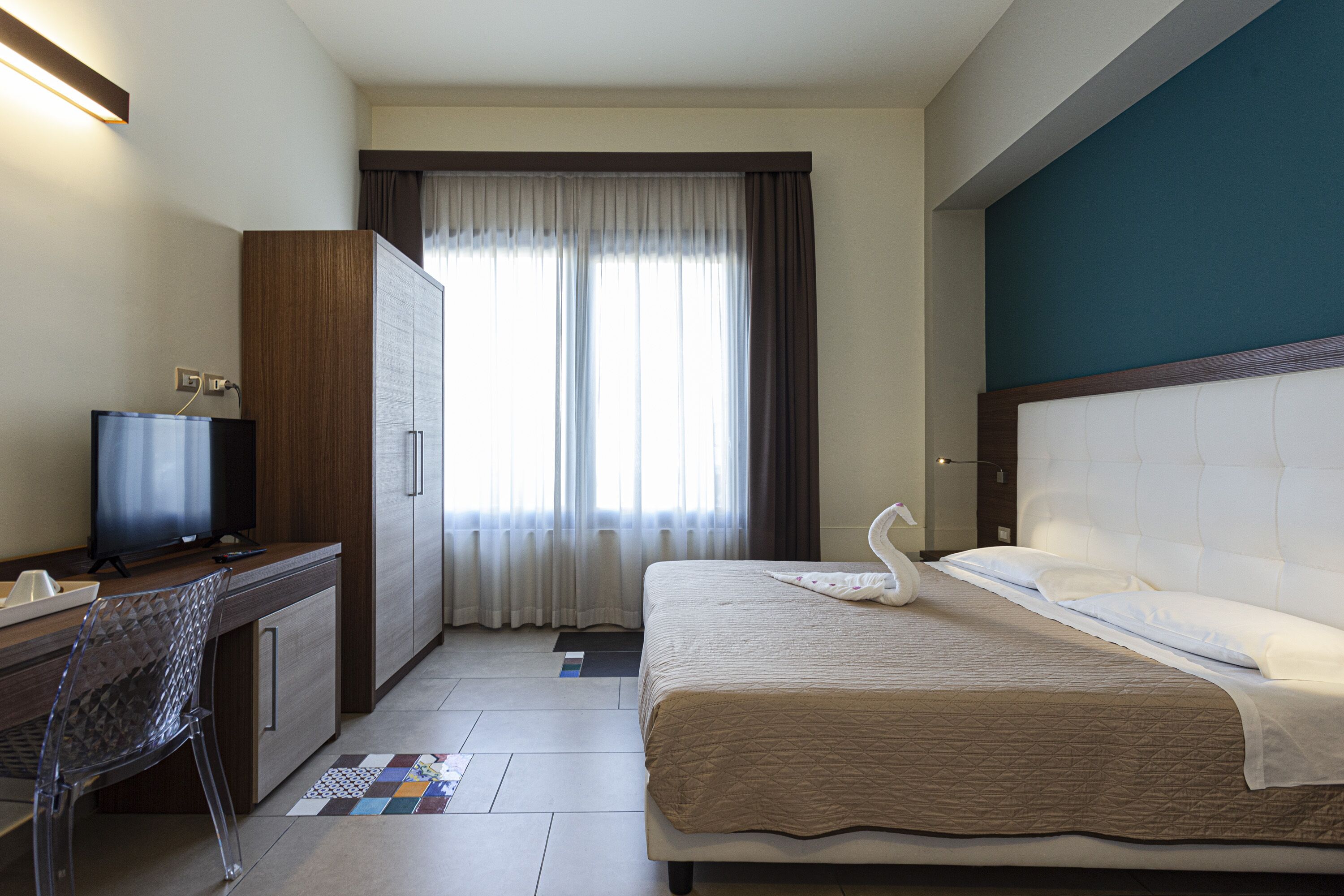A hotel room with a bed, a desk with a television, a wardrobe, and a window with curtains.