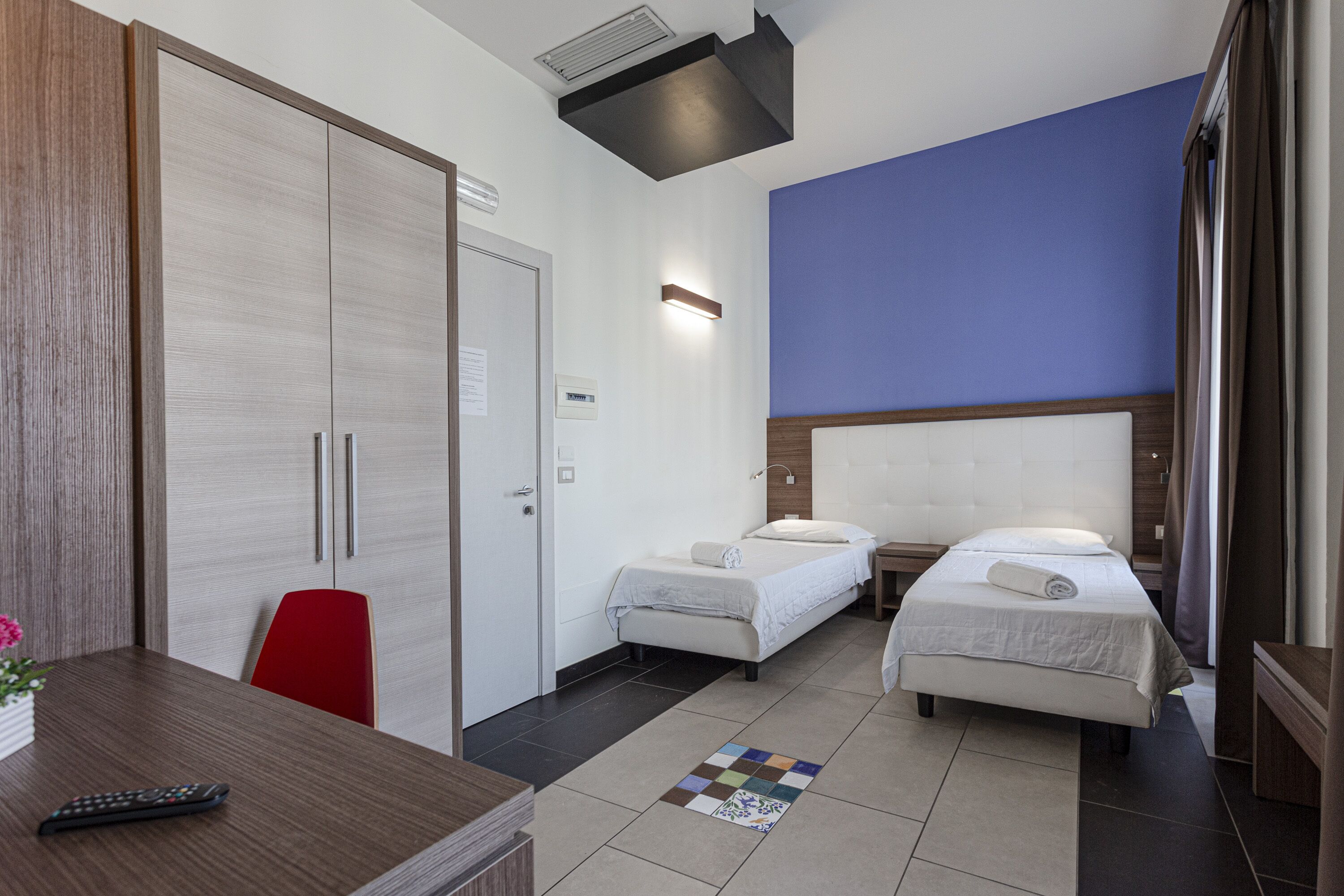 Habitación de hotel con dos camas individuales, un armario de madera, una silla roja, una mesita con un control remoto y un mostrador de recepción.