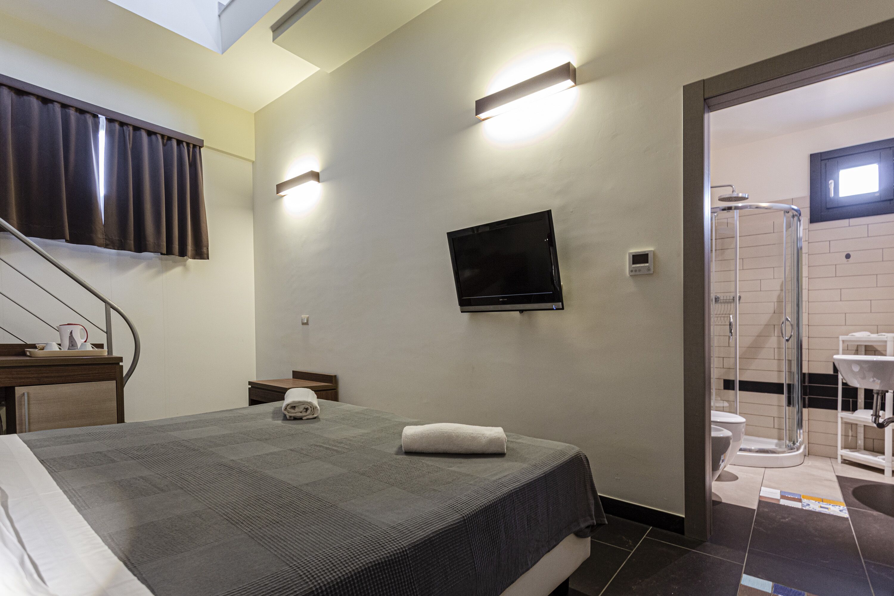 Une chambre d’hôtel équipée d’un lit, d’un téléviseur à écran plat fixé au mur, d’un espace douche et d’un petit bureau avec une tasse et un livre.