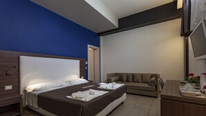 Habitación de hotel con una cama grande, un sofá, un escritorio con televisor y un baño visible a través de una puerta abierta.