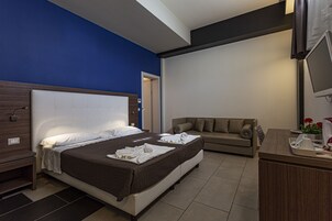 Habitación de hotel con una cama grande, un sofá, un escritorio con televisor y un baño visible a través de una puerta abierta.