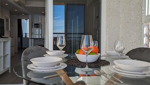 A dining area with a glass table set for four, surrounded by chairs. A bowl of fruit and wine glasses are on the table.